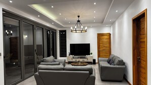 A modern living room with a large grey sofa, a smaller grey armchair, a TV, and a wooden door. - Tropical Elegance Haven - 4 bedrooms Villa on Mauritius’ Northwest Coast (Tombeau Bay)