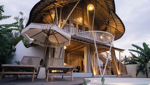 A modern, wooden structure with a curved roof and a balcony. There are wicker chairs and a table with a canopy. - Damuh Alam (Sukasada)