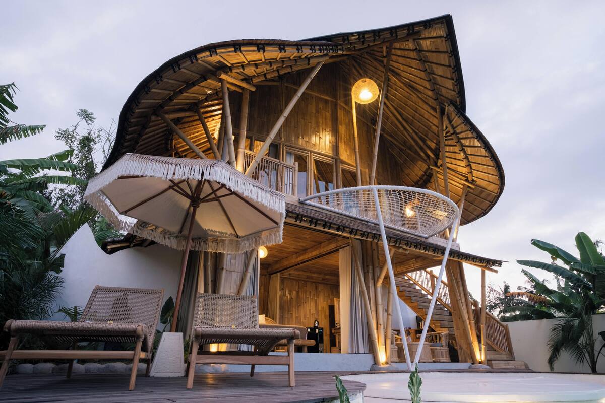 A modern, wooden structure with a curved roof and a balcony. There are wicker chairs and a table with a canopy.