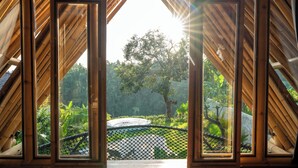 A view through a wooden-framed window with a metal railing, overlooking a garden with a large tree and a circular water feature. - Damuh Alam (Sukasada)