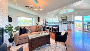 Ein geräumiges Wohnzimmer mit einer großen Couch, einem Couchtisch und einem schwarzen Sessel. Es gibt eine Küche mit einer großen Kochinsel und Barhocker. Der Raum verfügt über große Fenster und einen Blick ins Freie.