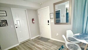 A room with two white doors, a fire extinguisher, a framed mirror, a small table with chairs, and a rug.