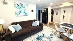 A living room with a brown sofa, a large abstract painting, a dining area with a white table and chairs, a refrigerator, and a coat rack.