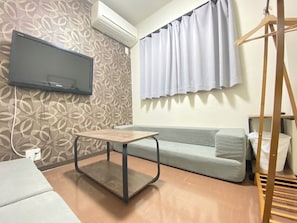 A room with a sofa, a table, a TV mounted on the wall, and a window with curtains.