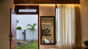 An interior space with a wooden door leading to an outdoor area, a seating arrangement, a wall-mounted mirror, and a window with curtains. - Jangal Resort - Sikar Road Jaipur (Jalsu)