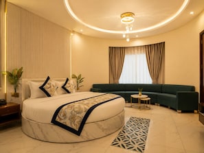 A modern bedroom with a large bed, a green sofa, a small round table, and a decorative rug. - Jangal Resort - Sikar Road Jaipur (Jalsu)