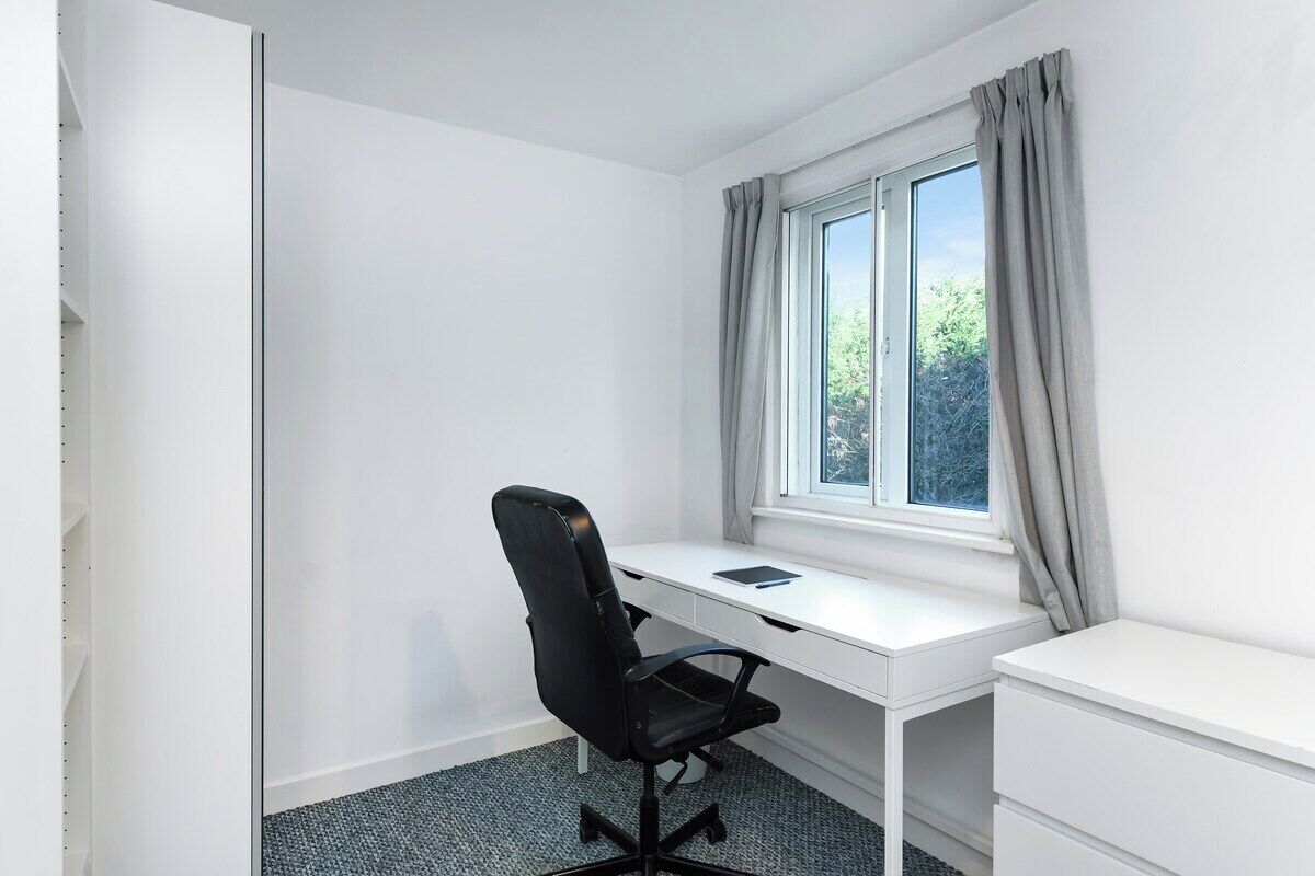 Un escritorio blanco con una silla de oficina negra, una ventana con cortinas y un armario blanco.