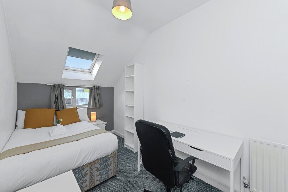 A hotel room with a bed, a desk, a chair, a radiator, and a window with curtains.