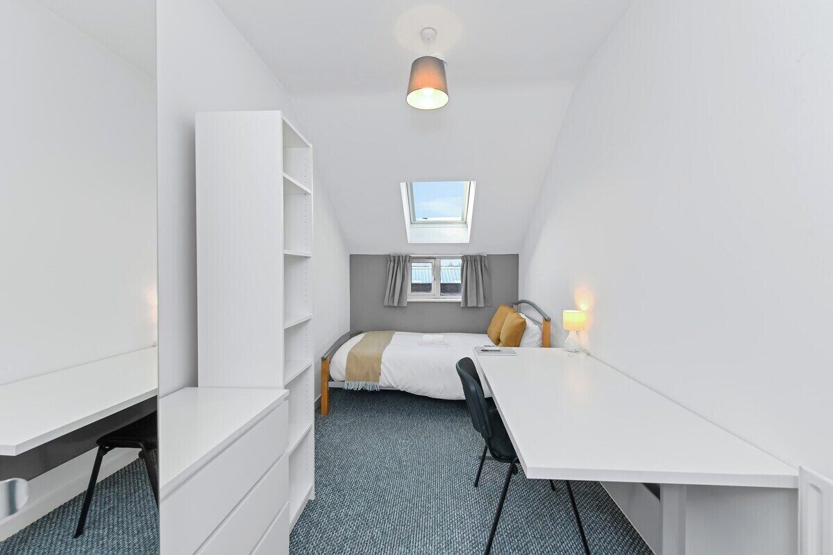Una habitación compacta con cama, escritorio, silla, estantería y tragaluz.
