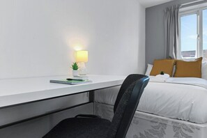 A hotel room with a desk, a chair, a bed with white and mustard pillows, a window with curtains, and a small plant on the desk. - Spacious 6-Bed Home Sleeps 12 Near Highfields Park (Nottingham)