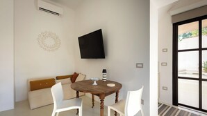 Ein modernes Wohnzimmer mit einem runden Holztisch, weißen Stühlen, einem beigefarbenen Sofa, einem flachbildfernseher an der Wand und einem großen Fenster mit Blick auf Grünflächen.