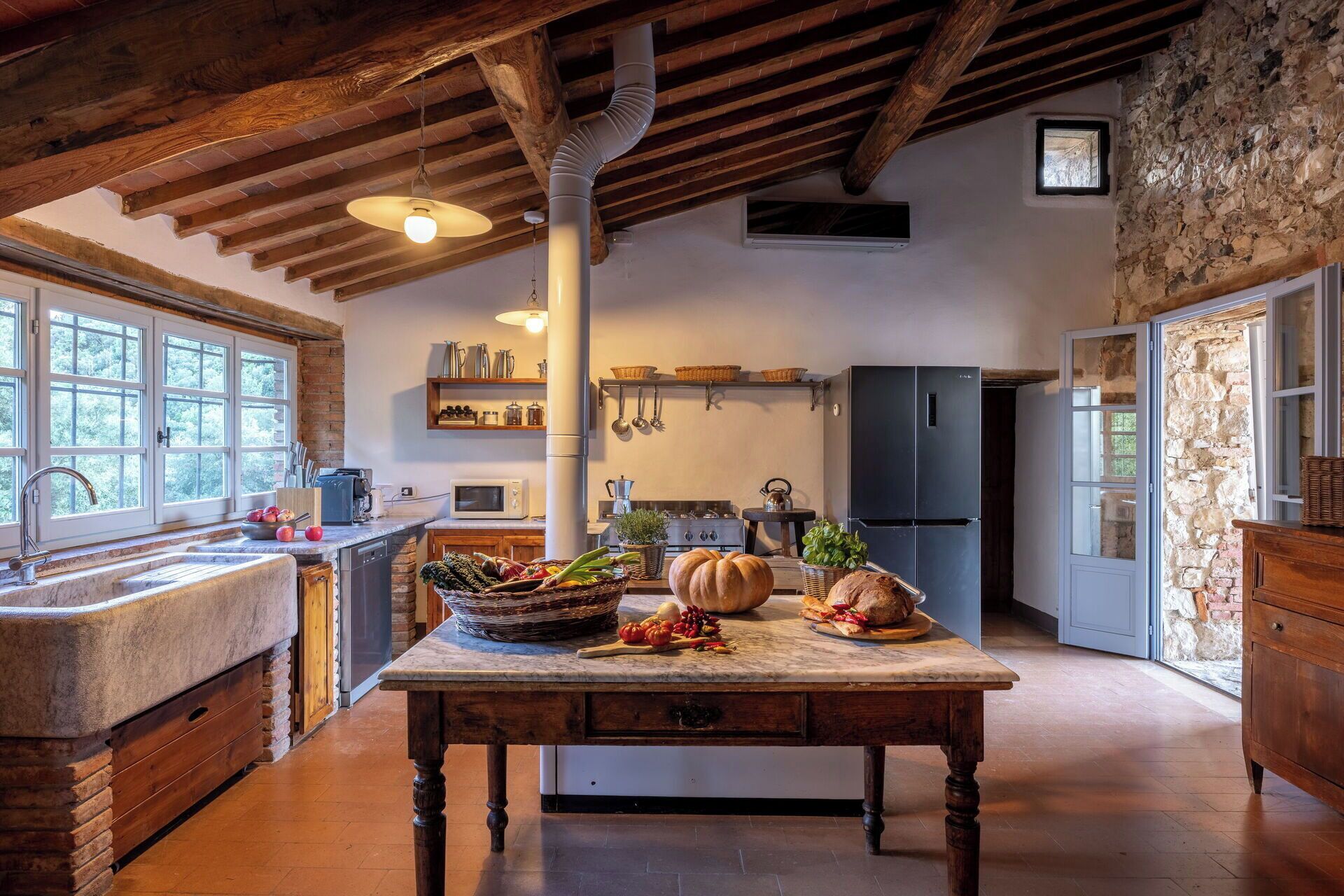 A rustic kitchen with exposed beams, a stone wall, and a wooden table with fresh produce.