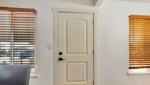 Una habitación con una puerta blanca, dos ventanas con persianas, una silla gris y suelo de madera.