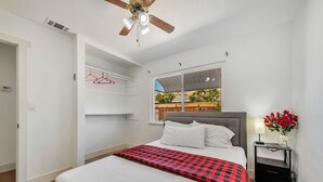 Un dormitorio con cama, ventilador de techo, ventana con vistas, mesita de noche con lámpara y estante de pared con perchas rojas.