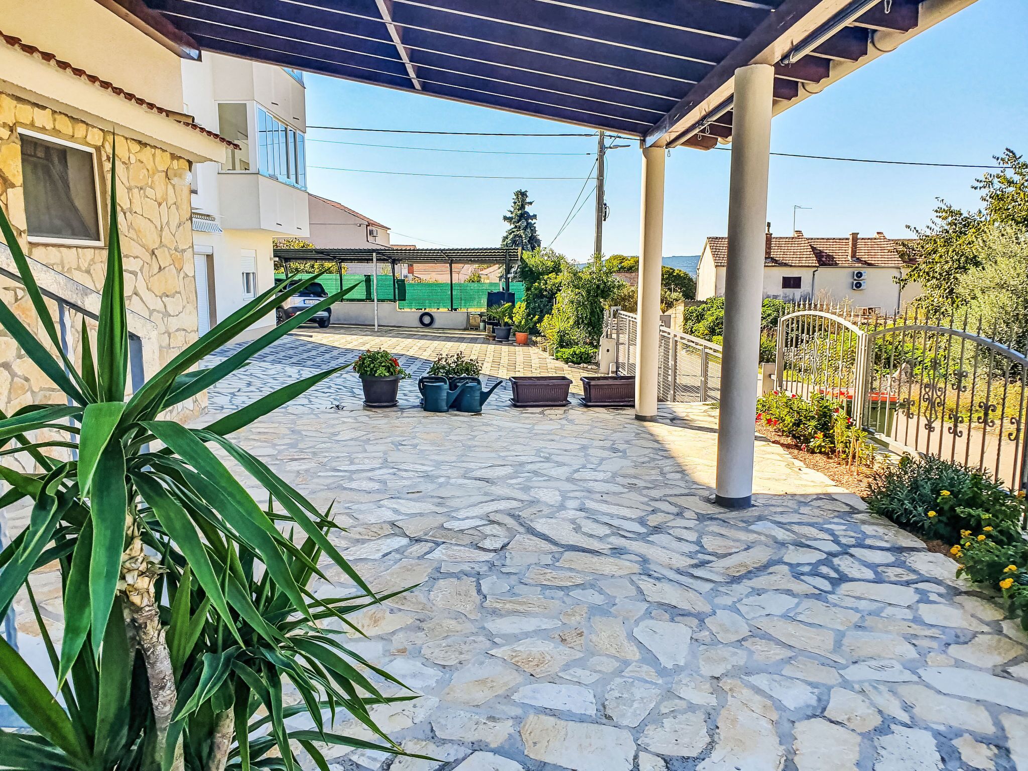 Un patio empedrado con zona de estar cubierta, plantas en macetas y una valla de metal.