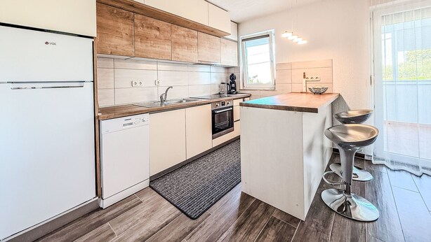 A compact kitchen with a built-in refrigerator, a sink, and a stove. There is a small island with stools and a window providing natural light. - Apartment Ana by Interhome (Sv. Filip i Jakov)