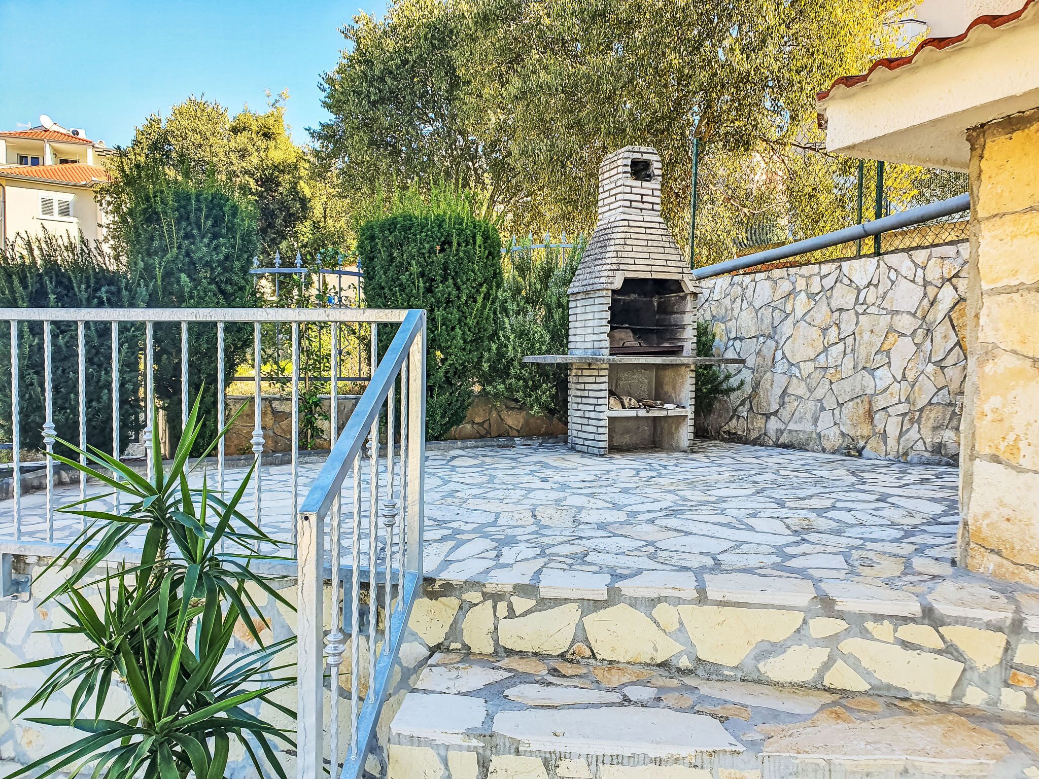 Un patio de piedra al aire libre con un horno de pizza de ladrillo, rodeado de vegetación y un muro de piedra.