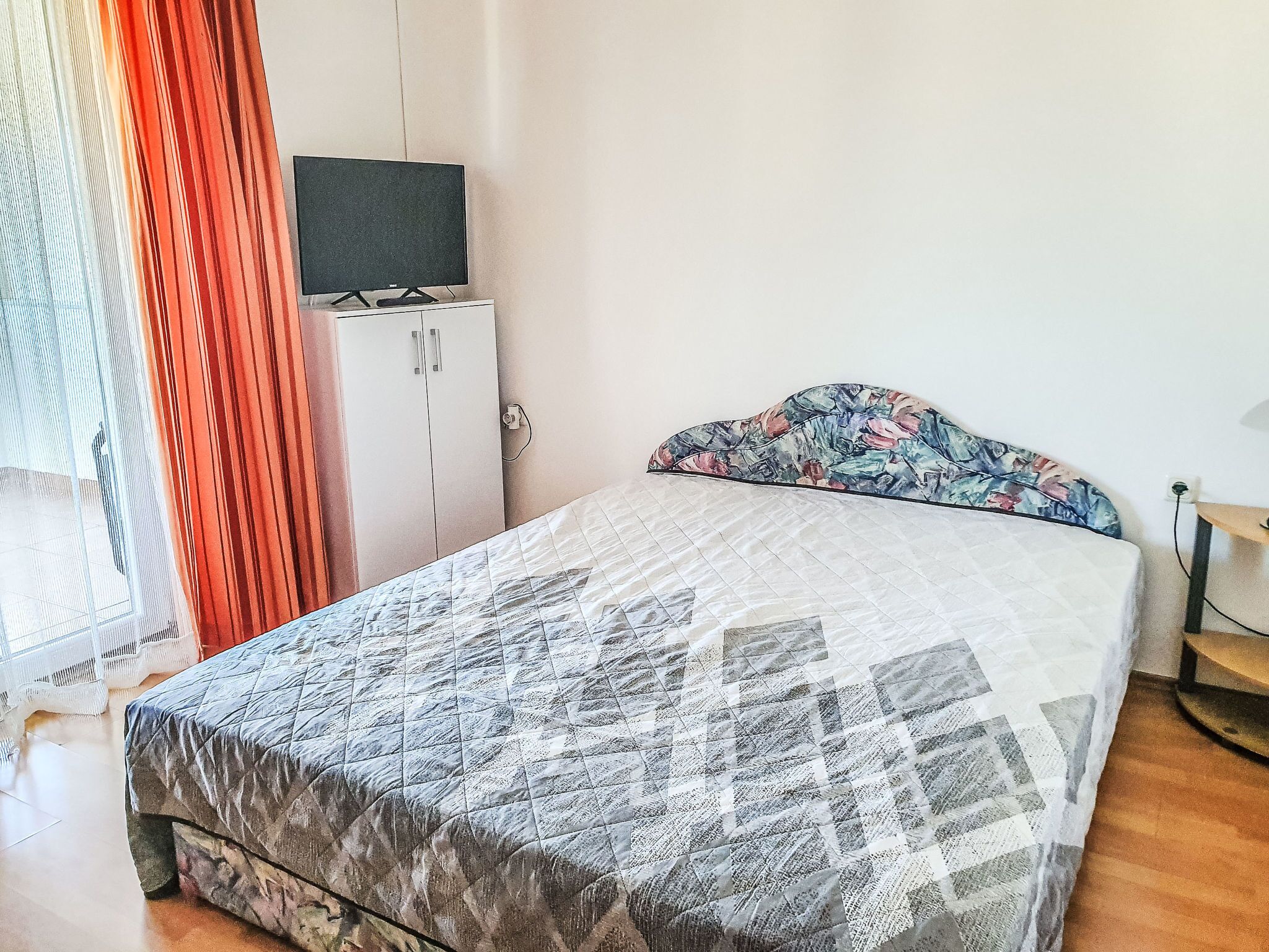 Quarto com cama, guarda-roupa, televisão em cima do guarda-roupa e janela com cortinas.