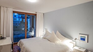 A hotel room with a bed, a bedside table, a nightstand, a lamp, and a view of buildings through a glass door. - Chesa Promulins by Interhome (Samedan)