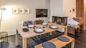 A modern dining area with a wooden table set for four, flanked by two cushioned benches. A television is mounted on the wall above the table, and a fireplace is visible in the background. - Chesa Promulins by Interhome (Samedan)