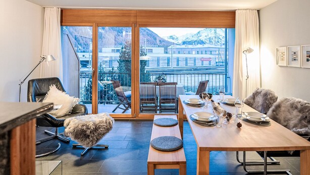 A modern dining area with a large table set for four, a sofa, and a chair. A large window offers a view of a snowy mountain and buildings. - Chesa Promulins by Interhome (Samedan)