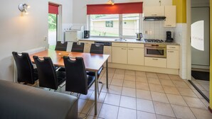 A compact kitchen with a dining area, featuring a stove, sink, and a window with a red valance. - 6-Person Comfort holiday home with Sauna. (Kootwijk)