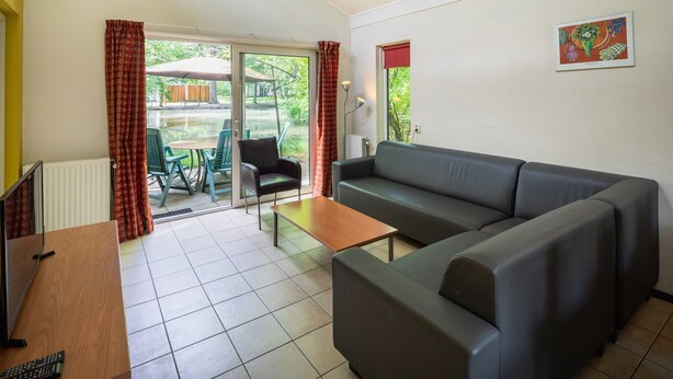 A living room with a sofa, a coffee table, and a TV. There is a sliding glass door leading to an outdoor area with a table and chairs. - 6-Person Comfort holiday home with Sauna. (Kootwijk)