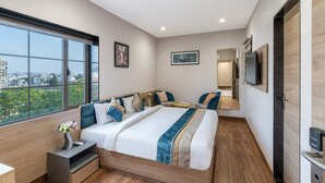 A modern hotel room with a large bed, a bedside table with a vase, a TV mounted on the wall, and a large window offering a city view. - The Hoften DiVi Hotel Aradhya Inn (Udaipur)