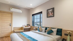 A modern hotel room with a large bed, a desk, a window with a city view, an air conditioning unit, and a painting on the wall. - The Hoften DiVi Hotel Aradhya Inn (Udaipur)