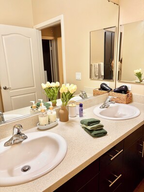 A bathroom with two sinks, a mirror, and a door. There are toiletries on the counter, including a bottle of lotion and a vase with flowers. - Ocean breeze retreat luxury 1B unit in Marina del Rey (Marina del Rey)