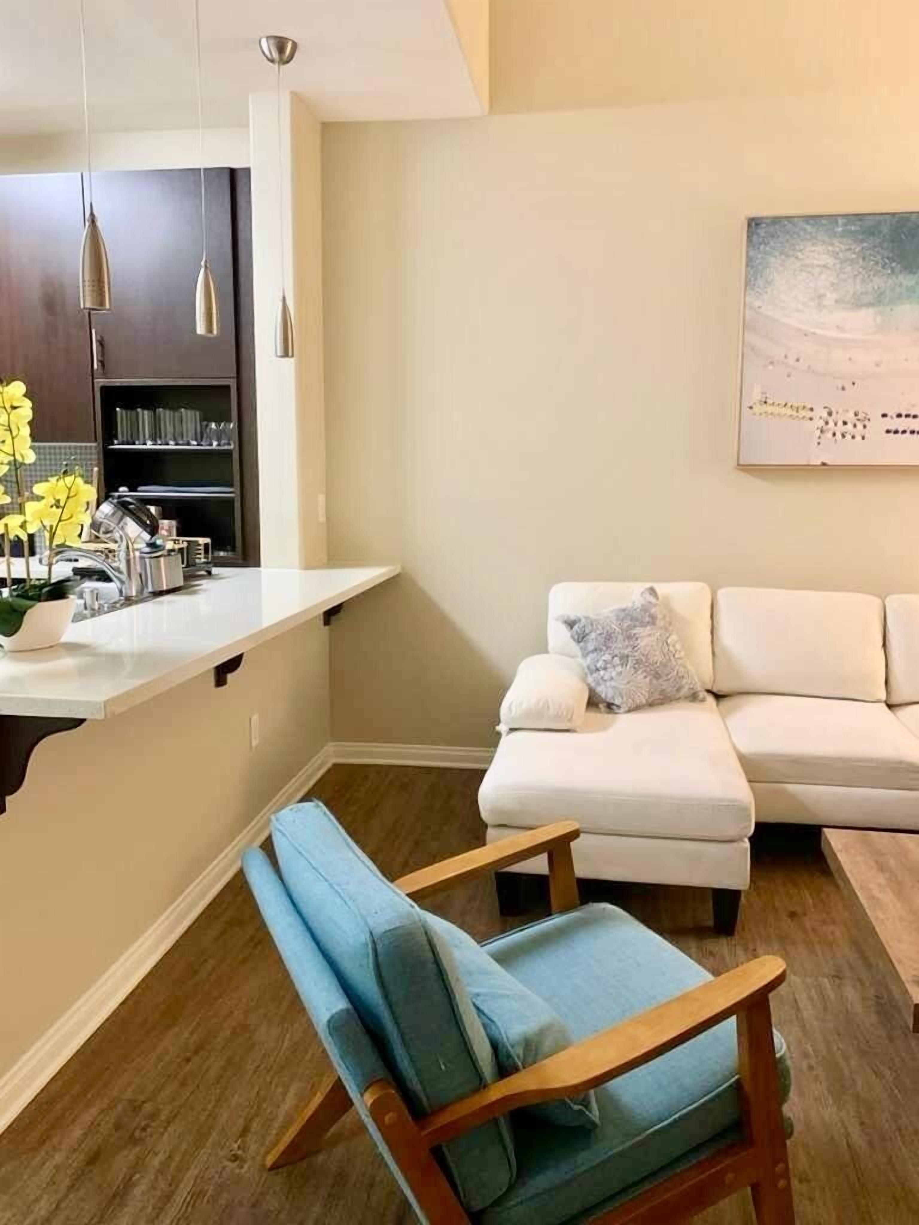 Una sala de estar moderna con un sofá blanco, una silla de madera y una zona de cocina con encimera blanca y luces colgantes.