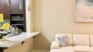 Una sala de estar moderna con un sofá blanco, una silla de madera y una zona de cocina con encimera blanca y luces colgantes.