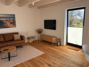 A modern living room with a wooden floor, a brown leather sofa, a round coffee table, a flat-screen TV mounted on the wall, a wooden bench, and a large window with a view of trees. - Gulfhof Wangermeer 2: A self-contained Tilda unit with a terrace and modern amenities - (Wangerland)