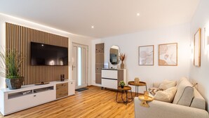 Ein modernes Wohnzimmer mit Flachbildfernseher, Holzboden, einer Couch und einer Essecke mit kleinem Tisch und Stühlen.