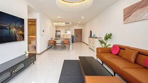 Ein modernes Wohnzimmer mit einem großen Fernseher, einem braunen Sofa, einer Chandelierleuchte und einem Essbereich. Im Hintergrund ist eine Küche zu sehen.