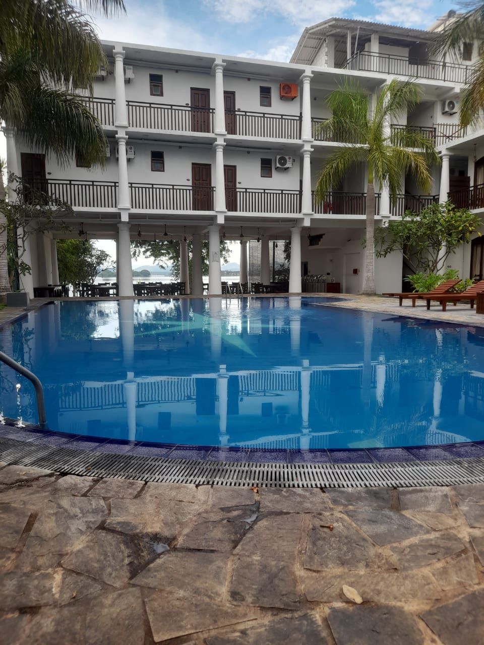 A pool area with a clear view of the pool, stone flooring, and a multi-story building with balconies and railings.
