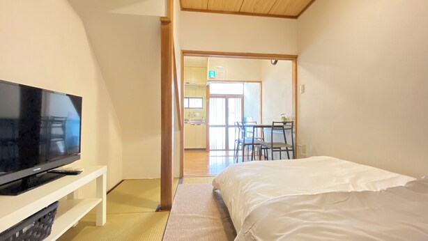 A bedroom with a bed, a TV on a stand, and a view of a dining area with chairs and a table. - AkiraHome Tsutenkaku West (Osaka)