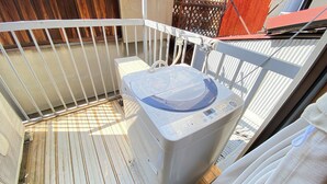 A compact, white washing machine on a balcony with a wooden floor and metal railing. - AkiraHome Tsutenkaku West (Osaka)