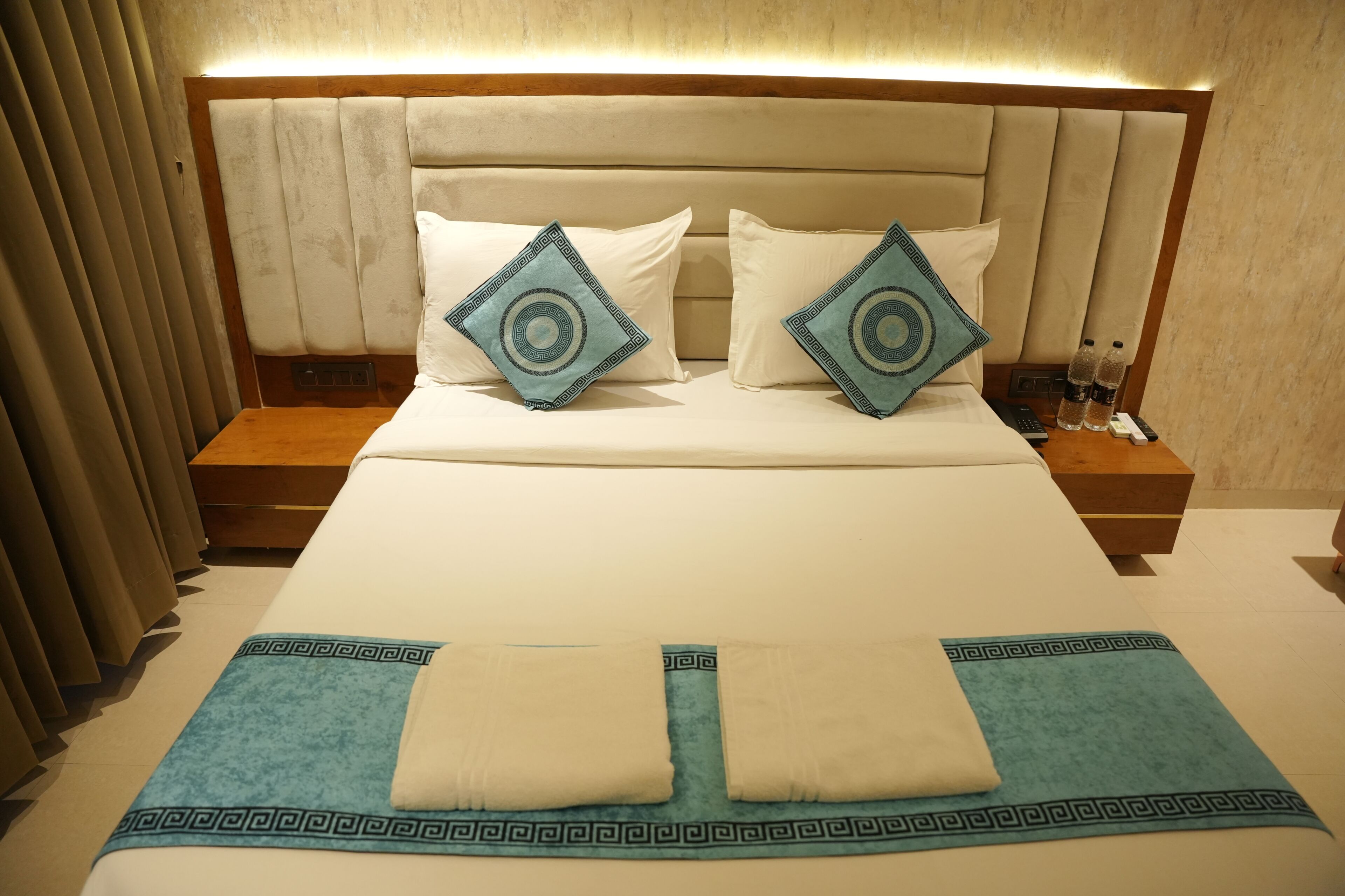 A neatly made bed with a headboard, two pillows, a blue throw blanket, and a bedside table with a remote control and water bottles.