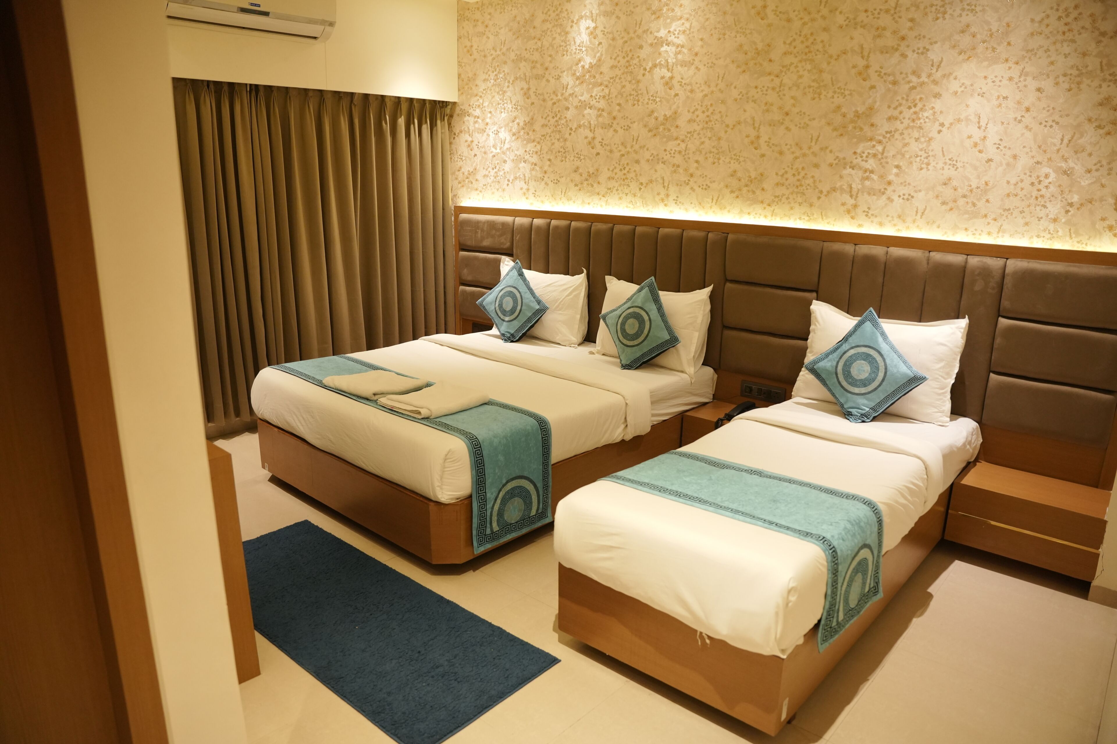 A hotel room with two beds, a large window with curtains, a wall air conditioner, and a blue carpet.