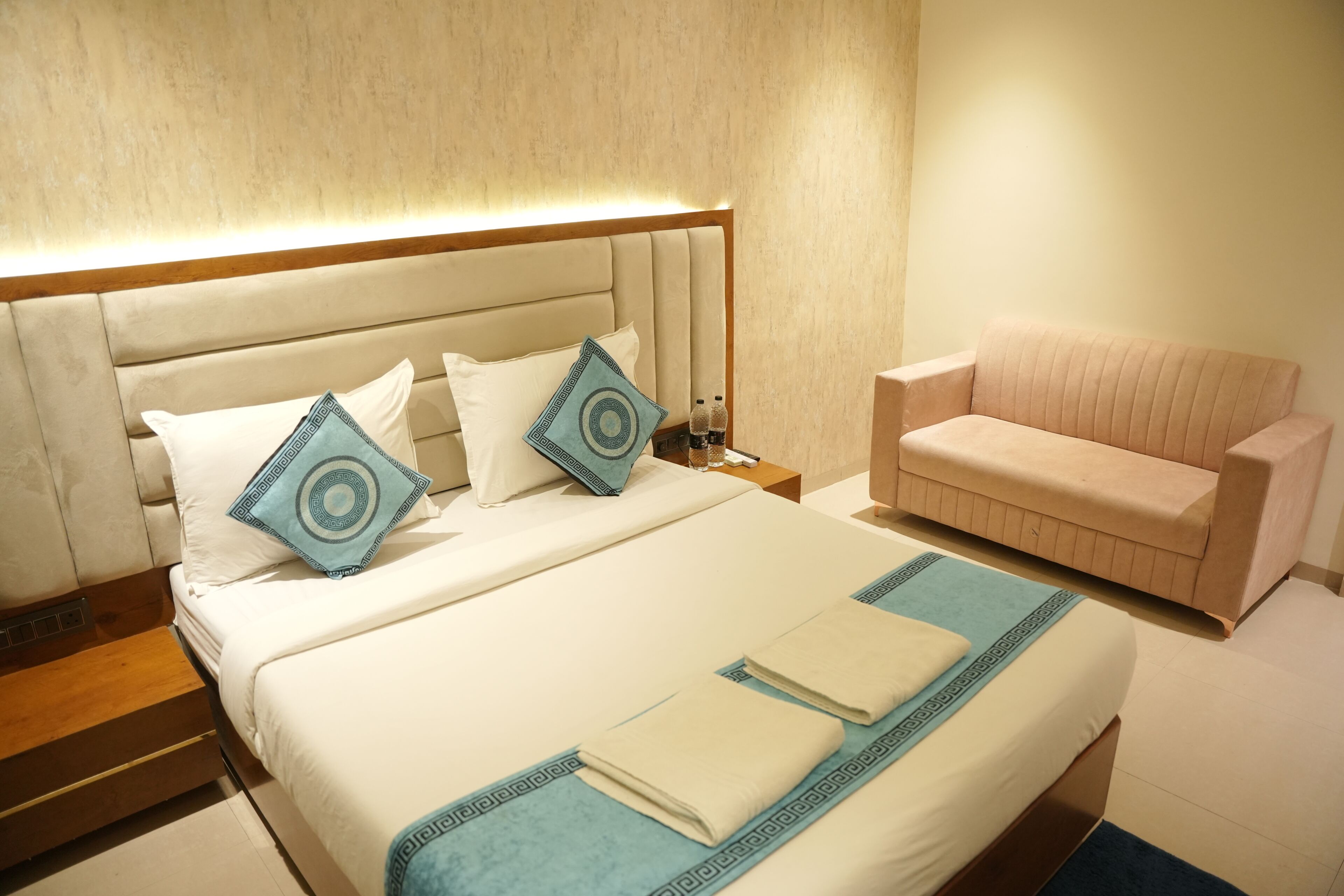 A hotel room with a bed, pillows, a nightstand, a sofa, and a wall-mounted lamp.