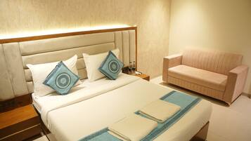A hotel room with a bed, pillows, a nightstand, a sofa, and a wall-mounted lamp.