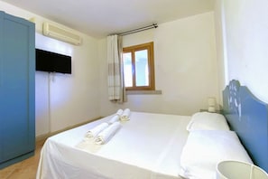 A hotel room with a bed, a TV mounted on the wall, an air conditioning unit, a window with curtains, and a small table with a lamp. - Charming two-room apartment of approximately 40 square meters with a 25 square meter covered veranda, located on the ground or first floor.It comprises a living room with a fully equipped kitchenette and a sofa bed, a bedroom with a double bed, and a bat (Porto Rotondo)