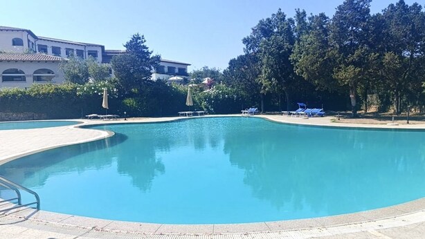 A large, clear swimming pool with a curved edge, surrounded by a tiled deck. There are lounge chairs and umbrellas along the poolside. - Charming two-room apartment of approximately 40 square meters with a 25 square meter covered veranda, located on the ground or first floor.It comprises a living room with a fully equipped kitchenette and a sofa bed, a bedroom with a double bed, and a bat (Porto Rotondo)
