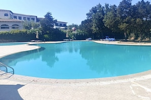 Una amplia piscina con borde curvo, rodeada por una terraza con baldosas. Se observan sillones de descanso y sombrillas a lo largo de la piscina.