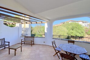 Un patio techado al aire libre con mobiliario de madera, incluyendo bancos y una mesa, y vistas a zonas verdes y edificios al fondo.