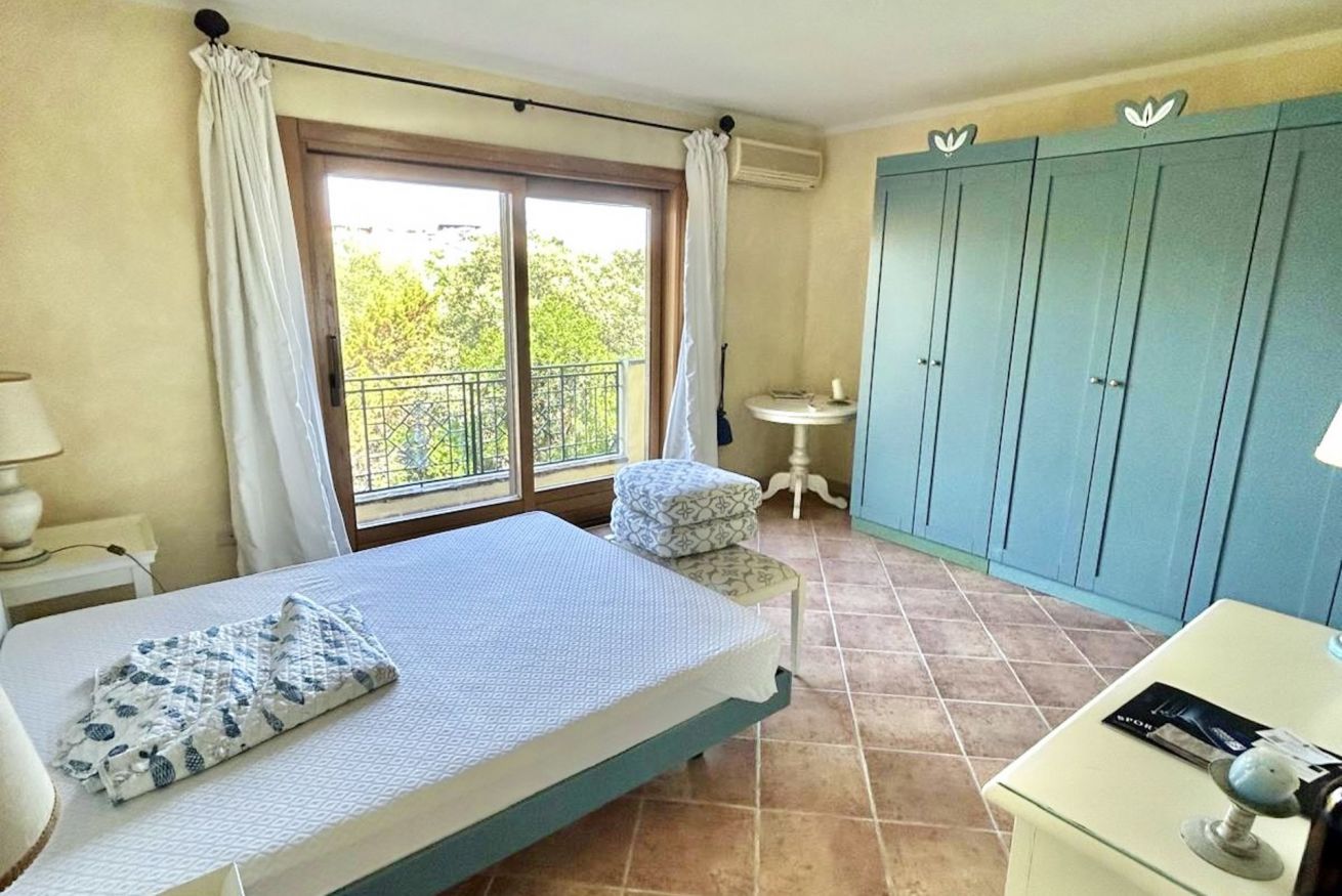 Ein Schlafzimmer mit einem Bett, einem kleinen Nachttisch, einem Sessel, einem Kleiderschrank und einem Fenster mit Vorhängen.