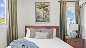 Ein ordentlich bezogenes Bett mit gemustertem Kopfteil, ein Nachttisch mit Lampe und ein gerahmtes Pflanzenbild an der Wand.