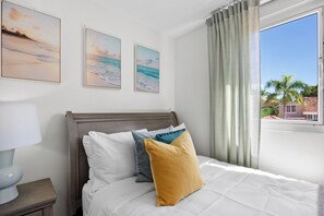 Ein Schlafzimmer mit einem Bett, Kissen, einem Nachttisch, einer Lampe und einem Fenster mit Vorhängen.