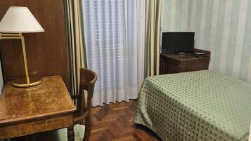 Una habitación de hotel con un escritorio de madera, una silla, una cama con edredón estampado en verde, un televisor sobre un mueble de madera y una ventana con cortinas.